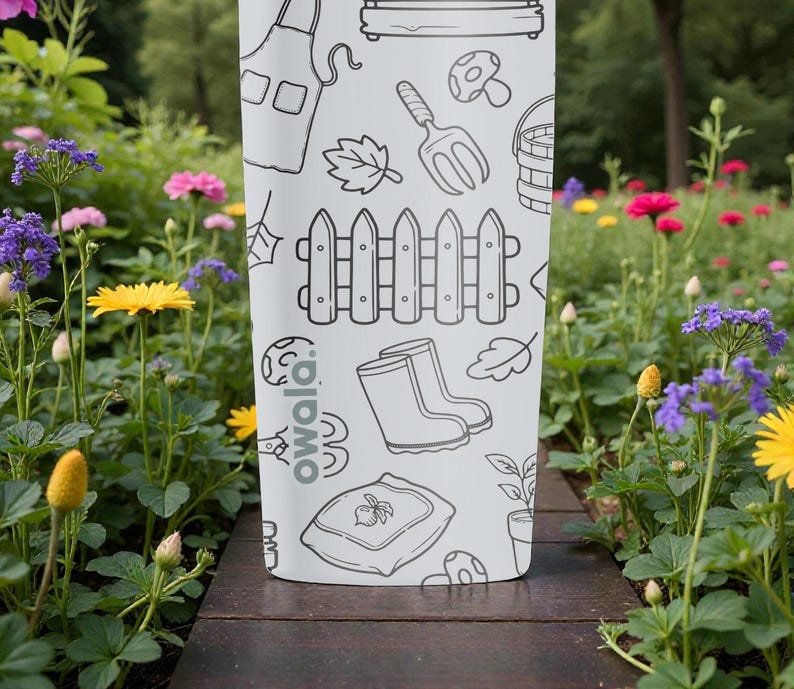 Puede incluir: Una botella de agua Owala blanca con un dise&ntilde;o de garabatos con tem&aacute;tica de jard&iacute;n. Las ilustraciones incluyen una valla, botas, una paleta y una seta. La botella est&aacute; sobre una superficie de madera, rodeada de flores.
