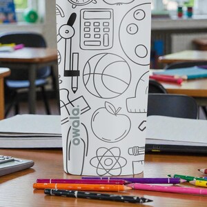 Op de afbeelding: Een witte waterfles met een zwart-wit schoolthema-ontwerp, inclusief een rekenmachine, passer, basketbal en appel. De fles heeft het woord "owala" erop gedrukt. Gekleurde potloden liggen verspreid op het houten bureau voor de fles.