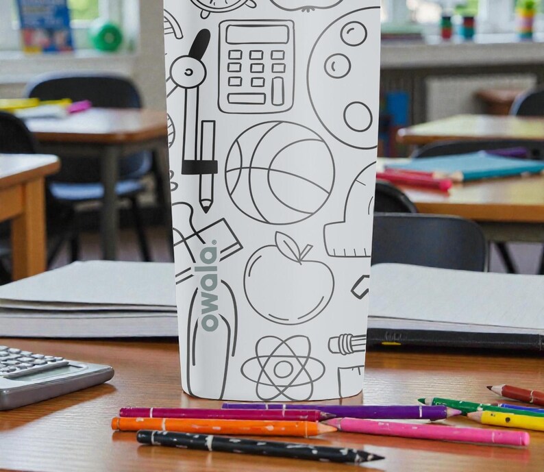 Op de afbeelding: Een witte waterfles met een zwart-wit schoolthema-ontwerp, inclusief een rekenmachine, passer, basketbal en appel. De fles heeft het woord "owala" erop gedrukt. Gekleurde potloden liggen verspreid op het houten bureau voor de fles.