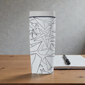 K&ouml;nnte beinhalten: Ein wei&szlig;er Reisebecher mit einem geometrischen Papierflieger-Design in Schwarz. Der Becher hat einen hellgrauen Deckel und einen dunkelgrauen Rand. Ein schwarzer Stift liegt auf einem wei&szlig;en Notizblock auf einer Holzoberfl&auml;che.