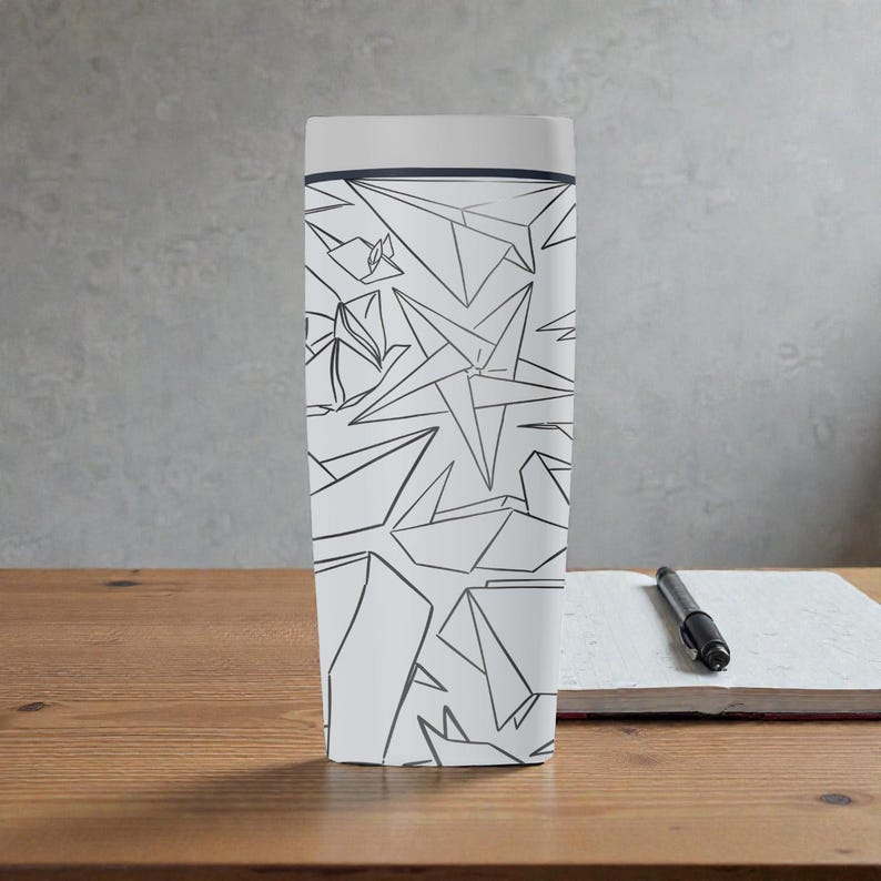 K&ouml;nnte beinhalten: Ein wei&szlig;er Reisebecher mit einem geometrischen Papierflieger-Design in Schwarz. Der Becher hat einen hellgrauen Deckel und einen dunkelgrauen Rand. Ein schwarzer Stift liegt auf einem wei&szlig;en Notizblock auf einer Holzoberfl&auml;che.
