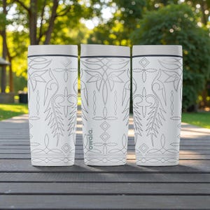 May include: Three white insulated tumblers with a decorative stitched design. The tumblers have a grey lid and a black trim. The design features floral and geometric patterns. The brand name "owala" is printed on the side of the tumblers.