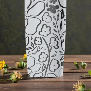 May include: White tumbler with a black floral design of flowers, leaves, and a butterfly. The tumbler is on a wooden surface with yellow flowers and green buds scattered around it.
