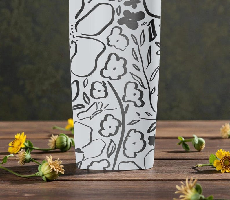 May include: White tumbler with a black floral design of flowers, leaves, and a butterfly. The tumbler is on a wooden surface with yellow flowers and green buds scattered around it.