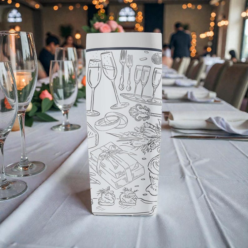 May include: A white gift box with black line drawings of champagne glasses, forks, rings, and a wrapped gift. The box is on a white tablecloth with wine glasses and a floral arrangement in the background.