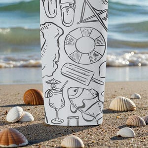 Op de afbeelding: Een witte beker met een zwarte lijntekening van strandthema's, waaronder een reddingsboei, een ijsje en een vis. De beker staat op een zandstrand met schelpen.