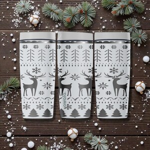 May include: Three white insulated tumblers with a festive winter design. Each tumbler features a pattern of reindeer, snowflakes, and trees in gray. The tumblers are set against a dark wood background with holiday decorations.
