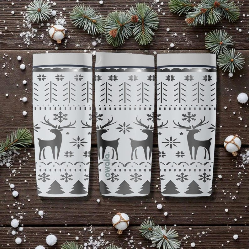 May include: Three white insulated tumblers with a festive winter design. Each tumbler features a pattern of reindeer, snowflakes, and trees in gray. The tumblers are set against a dark wood background with holiday decorations.