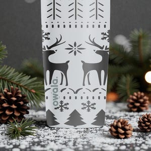 May include: A white Owala water bottle with a black holiday design featuring reindeer, snowflakes, and trees. The bottle is surrounded by pine needles, pine cones, and artificial snow, creating a festive winter scene.