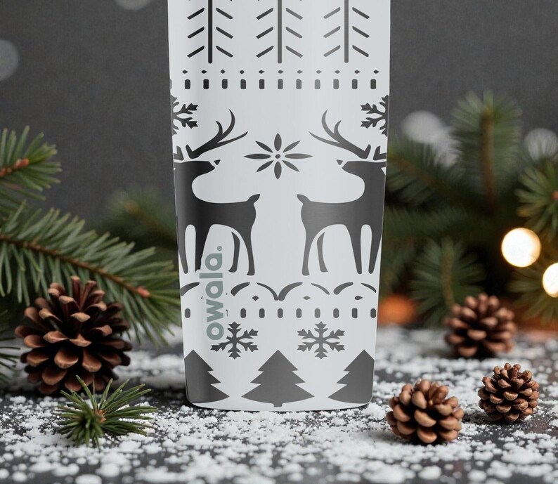 May include: A white Owala water bottle with a black holiday design featuring reindeer, snowflakes, and trees. The bottle is surrounded by pine needles, pine cones, and artificial snow, creating a festive winter scene.