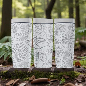 Op de afbeelding: Drie witte geïsoleerde waterflessen met een zwart-wit monstera bladpatroon. De flessen hebben een grijze deksel en een zwarte rand. Het woord "owala!" staat op een van de flessen.