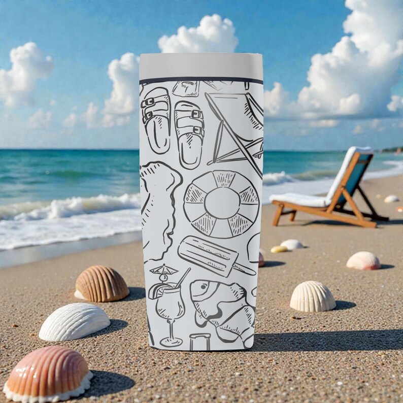 Op de afbeelding: Een wit en grijze reismok met een strandthema-ontwerp met lijntekeningen van sandalen, een strandstoel, een reddingsboei en andere zomericonen. De mok staat op een zandstrand met schelpen en de oceaan op de achtergrond.