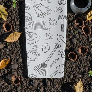 Puede incluir: Una toalla blanca con tem&aacute;tica de jard&iacute;n con ilustraciones en negro de herramientas y elementos de jardiner&iacute;a, incluyendo una regadera, un rastrillo, guantes y hojas. La toalla est&aacute; rodeada de tierra, hojas y c&aacute;scaras de nueces.