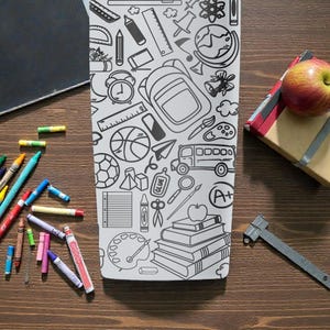 May include: A white tumbler with a black school-themed doodle design sits on a wooden surface, surrounded by crayons, a chalkboard, a wrapped gift, an apple, and a metal ruler. The tumbler features illustrations of school supplies.