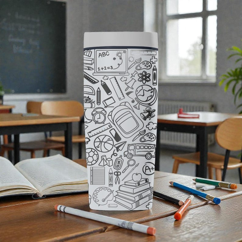May include: A white and gray travel mug with a school-themed doodle design. The design includes images of a laptop, ruler, backpack, books, and other school supplies. The mug is sitting on a wooden desk with pencils and an open book.