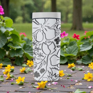 May include: A white, rectangular tumbler with a black floral design. The tumbler has a light grey top. The floral pattern includes roses, daisies, and other stylized flowers. The background features flowers and greenery.