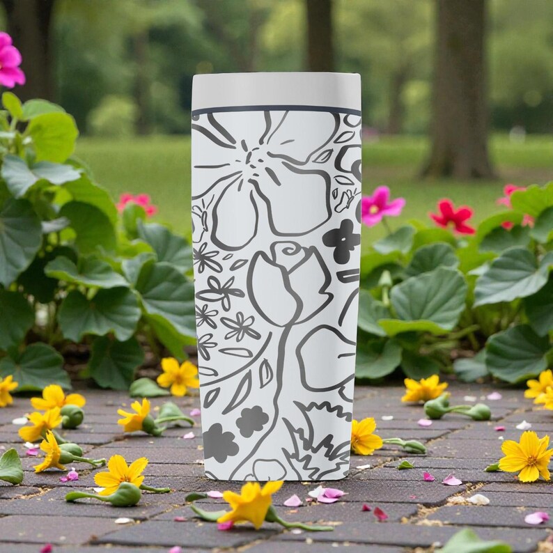 May include: A white, rectangular tumbler with a black floral design. The tumbler has a light grey top. The floral pattern includes roses, daisies, and other stylized flowers. The background features flowers and greenery.