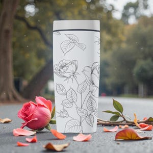 Op de afbeelding: Een witte reismok met een zwarte lijntekening van rozen en bladeren. Een roze roos en verspreide bloemblaadjes staan op de voorgrond. De mok heeft een wit deksel en een zwarte band aan de bovenkant.
