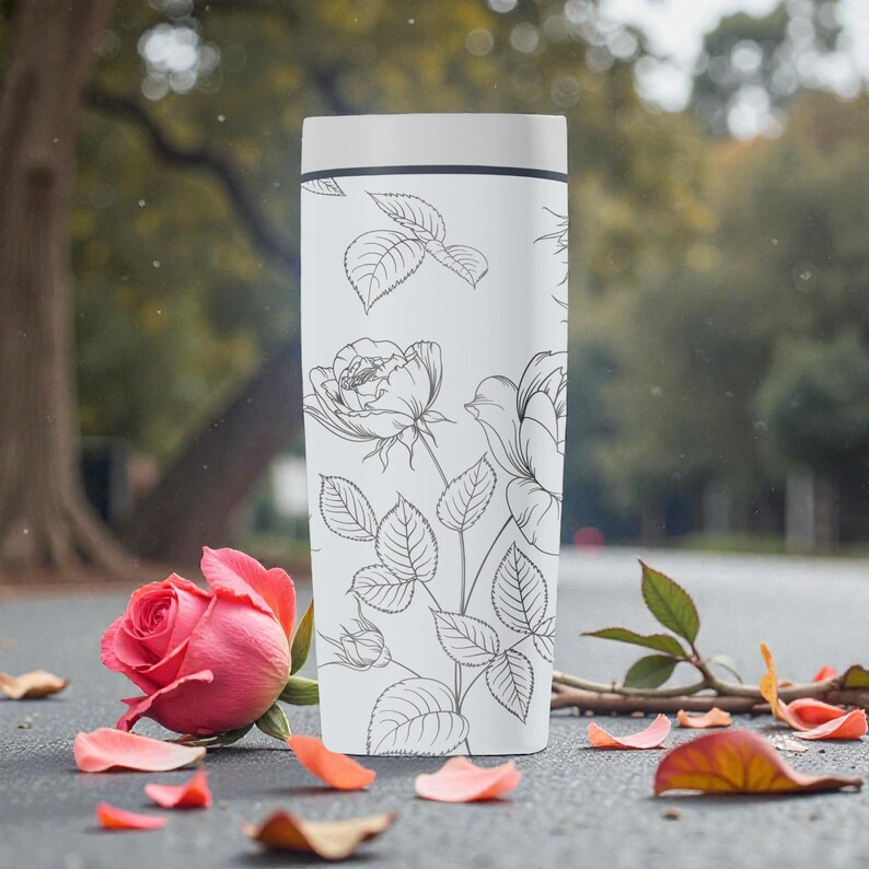 Op de afbeelding: Een witte reismok met een zwarte lijntekening van rozen en bladeren. Een roze roos en verspreide bloemblaadjes staan op de voorgrond. De mok heeft een wit deksel en een zwarte band aan de bovenkant.