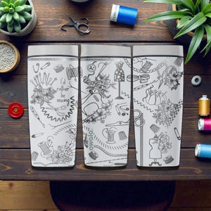 May include: Three white travel mugs with black line art of sewing notions and floral designs. One mug has the text "I'd rather be sewing." The mugs are on a dark wood surface with sewing supplies and plants.