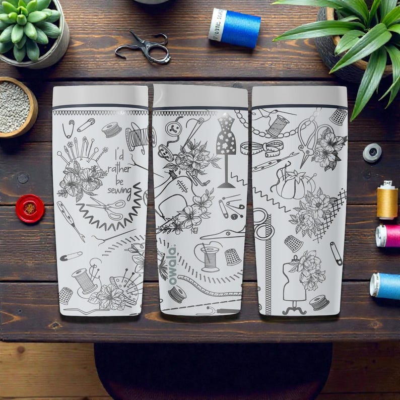 May include: Three white travel mugs with black line art of sewing notions and floral designs. One mug has the text "I'd rather be sewing." The mugs are on a dark wood surface with sewing supplies and plants.