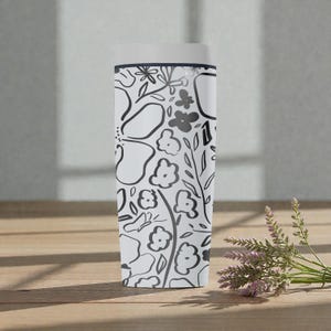 May include: A white travel mug featuring a black floral design. The pattern includes stylized flowers, leaves, and other botanical elements. The mug has a white lid and is approximately 20 cm tall. The mug is on a wooden surface.