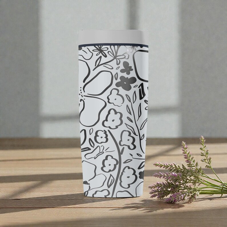 May include: A white travel mug featuring a black floral design. The pattern includes stylized flowers, leaves, and other botanical elements. The mug has a white lid and is approximately 20 cm tall. The mug is on a wooden surface.