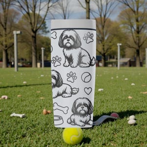 May include: A white tumbler featuring a Shih Tzu dog design, paw prints, a bone, a bowl, a tennis ball, and a heart. The tumbler is cylindrical and stands upright on a grassy surface. The background shows a park setting with trees and a green lawn.