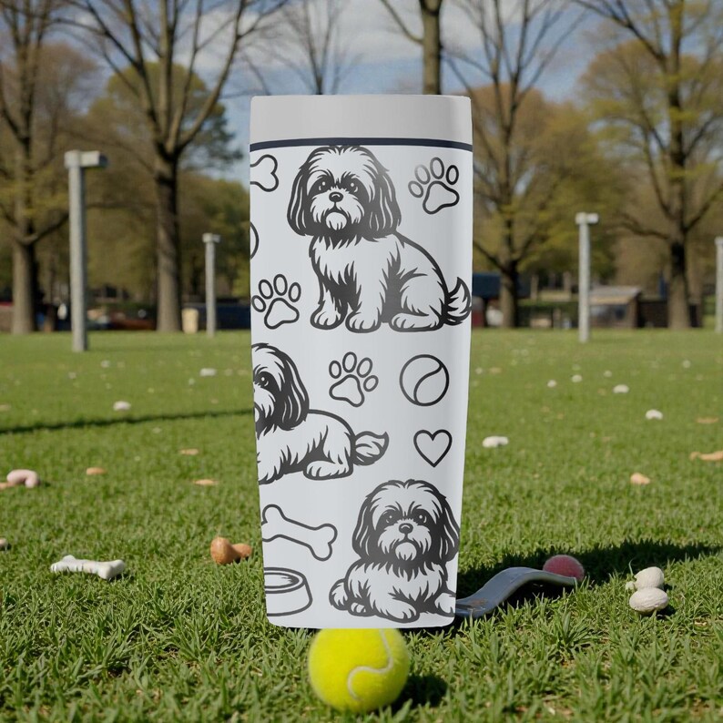 May include: A white tumbler featuring a Shih Tzu dog design, paw prints, a bone, a bowl, a tennis ball, and a heart. The tumbler is cylindrical and stands upright on a grassy surface. The background shows a park setting with trees and a green lawn.