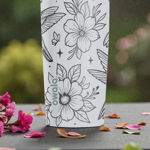 Puede incluir: Una botella de agua blanca Owala con un dise&ntilde;o floral y colibr&iacute; negro. El dise&ntilde;o incluye flores, colibr&iacute;es, mariposas y estrellas. La botella est&aacute; sobre una superficie oscura con p&eacute;talos de flores rosas.