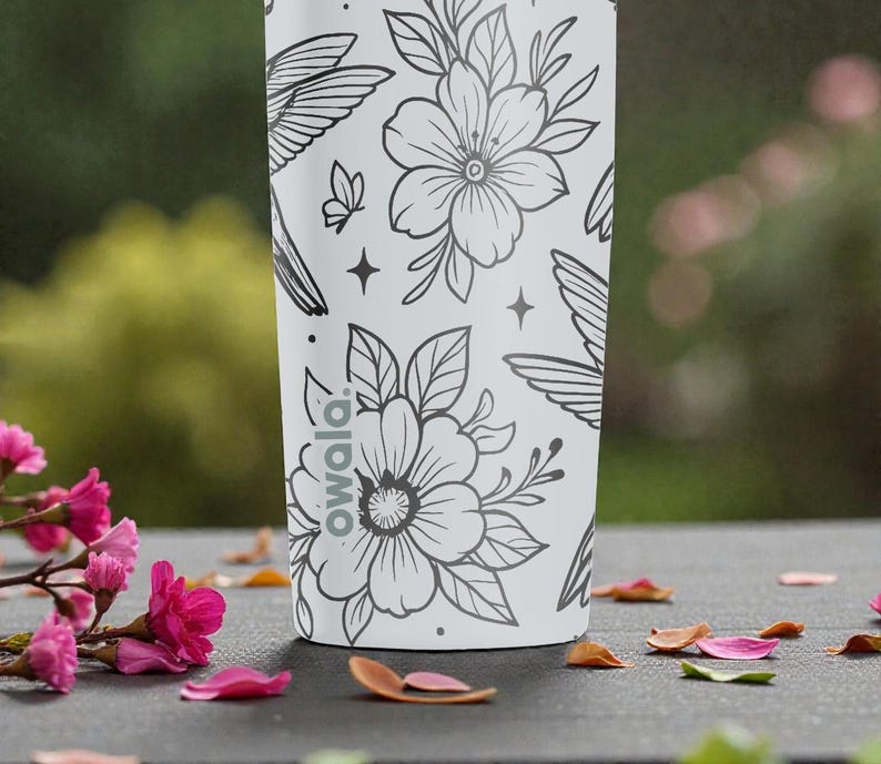 Puede incluir: Una botella de agua blanca Owala con un dise&ntilde;o floral y colibr&iacute; negro. El dise&ntilde;o incluye flores, colibr&iacute;es, mariposas y estrellas. La botella est&aacute; sobre una superficie oscura con p&eacute;talos de flores rosas.