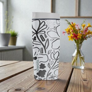 May include: White Owala water bottle with a black floral design. The bottle has a white lid and a dark grey band. The bottle is on a wooden table. A vase of flowers is in the background.
