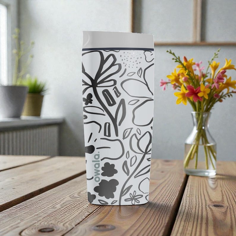 May include: White Owala water bottle with a black floral design. The bottle has a white lid and a dark grey band. The bottle is on a wooden table. A vase of flowers is in the background.