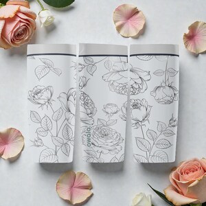 Op de afbeelding: Drie witte waterflessen met een bloemenrozenontwerp. De flessen hebben een grijze bovenkant en zijn versierd met zwarte lijntekeningen van rozen en bladeren. Het woord "owala" staat op de zijkant van een fles. Roze rozenblaadjes en rozen zijn rond de flessen verspreid.
