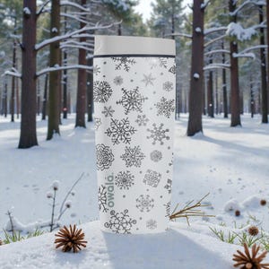 Puede incluir: Taza de viaje blanca con tapa gris y un patr&oacute;n de copos de nieve negros. La palabra "owala" est&aacute; impresa en el lateral. La taza cil&iacute;ndrica parece estar aislada. El fondo es un bosque nevado.