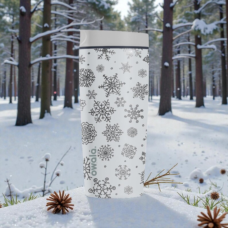 Puede incluir: Taza de viaje blanca con tapa gris y un patr&oacute;n de copos de nieve negros. La palabra "owala" est&aacute; impresa en el lateral. La taza cil&iacute;ndrica parece estar aislada. El fondo es un bosque nevado.