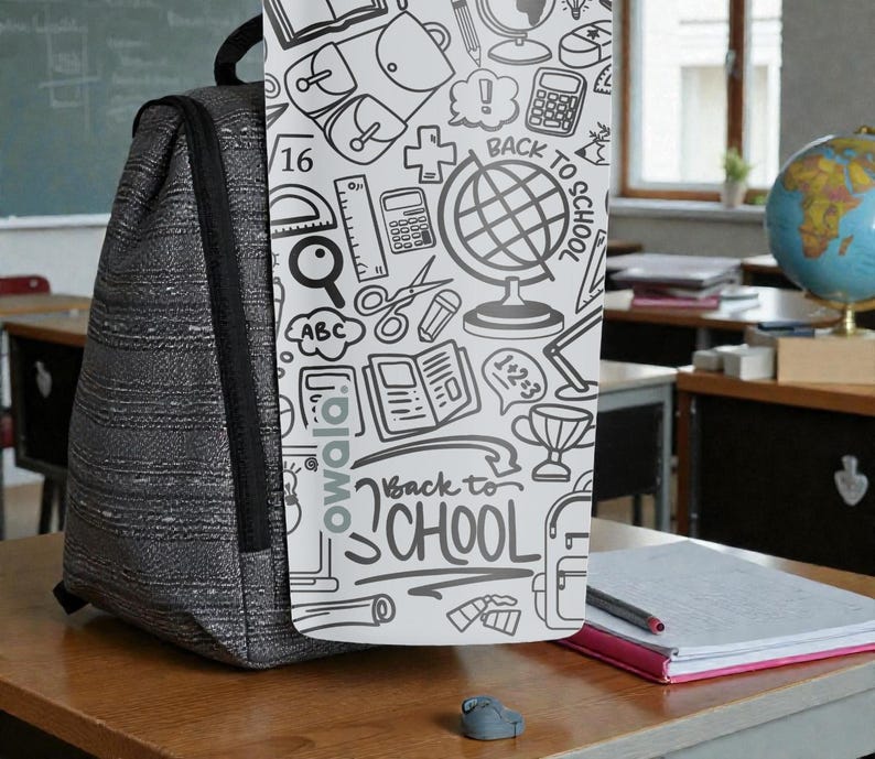May include: A gray backpack with a black strap stands on a wooden desk. A white panel with school-themed doodles and the text "Back to School" is attached to the backpack. A globe and notebook are visible in the background.
