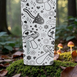 Peut inclure: Une gourde blanche avec un motif noir r&eacute;p&eacute;t&eacute; de champignons, de c&oelig;urs, d'&eacute;toiles et de fleurs. La gourde porte le mot "owala". La gourde est plac&eacute;e sur un fond de for&ecirc;t flou avec des champignons.