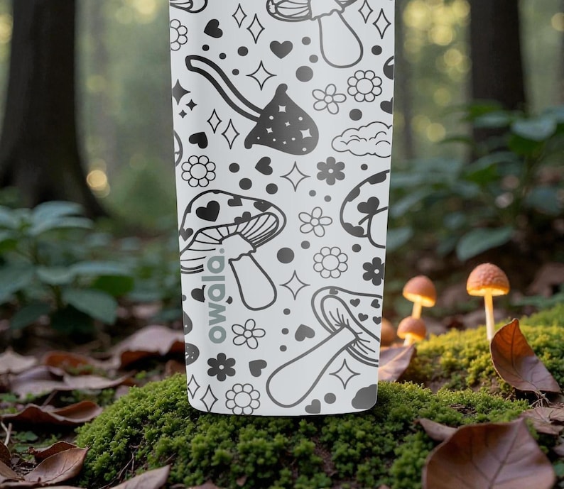 Peut inclure: Une gourde blanche avec un motif noir r&eacute;p&eacute;t&eacute; de champignons, de c&oelig;urs, d'&eacute;toiles et de fleurs. La gourde porte le mot "owala". La gourde est plac&eacute;e sur un fond de for&ecirc;t flou avec des champignons.