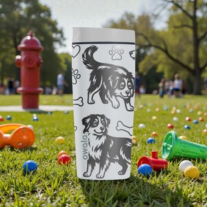 Puede incluir: Una botella de agua Owala blanca con ilustraciones en blanco y negro de perros, huellas de patas y huesos. La botella tiene una tapa blanca. El fondo muestra un parque con juguetes y una boca de incendios roja.