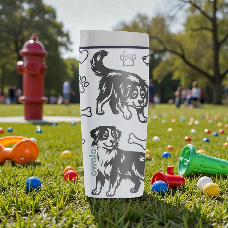 Puede incluir: Una botella de agua Owala blanca con ilustraciones en blanco y negro de perros, huellas de patas y huesos. La botella tiene una tapa blanca. El fondo muestra un parque con juguetes y una boca de incendios roja.