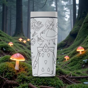 May include: A white travel mug featuring black line art illustrations of characters from Alice in Wonderland. The design includes the Cheshire Cat, Alice, the Queen of Hearts, and other characters. The mug has a light gray lid.