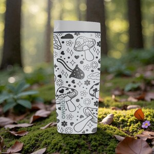 Peut inclure: Un mug de voyage blanc avec un motif de champignons noir et blanc. Le mug a un couvercle blanc et un bord noir. Le motif comprend des champignons, des fleurs, des c&oelig;urs et des diamants. La marque "owala!" est imprim&eacute;e sur le c&ocirc;t&eacute;.
