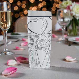 May include: A white Owala water bottle with black line art of a heart, flowers, and other designs. The bottle is on a table with rose petals and glasses of champagne. The background has blurred lights.