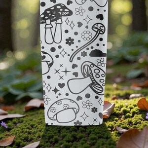 Peut inclure: Un gobelet blanc avec un motif de champignons noir et blanc. Le motif comprend diverses illustrations de champignons, des fleurs, des c&oelig;urs et des &eacute;toiles. Le gobelet cylindrique est pos&eacute; sur un fond naturel flou.
