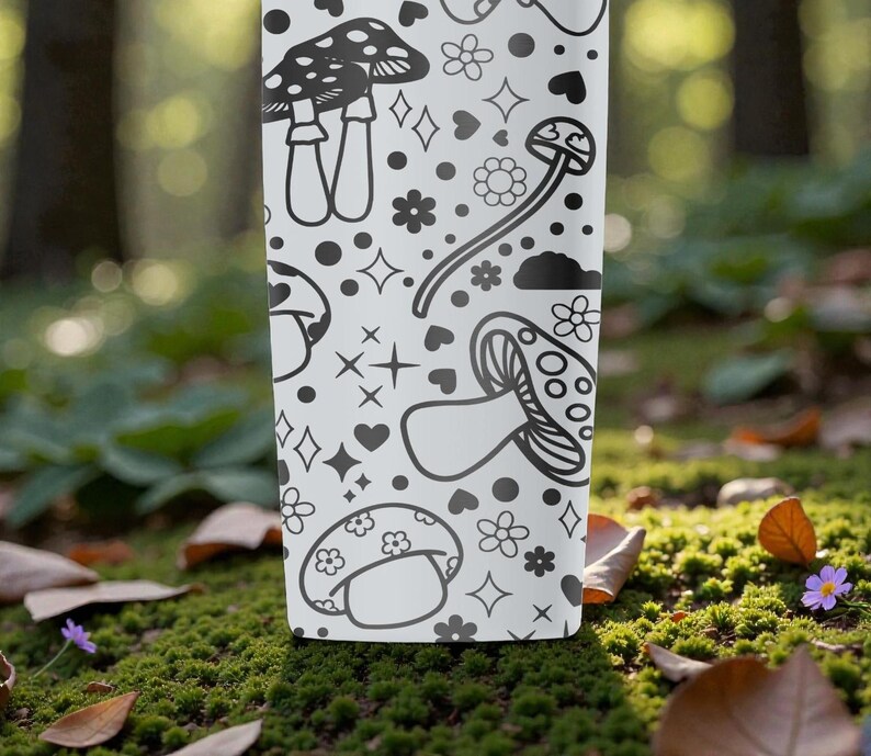Peut inclure: Un gobelet blanc avec un motif de champignons noir et blanc. Le motif comprend diverses illustrations de champignons, des fleurs, des c&oelig;urs et des &eacute;toiles. Le gobelet cylindrique est pos&eacute; sur un fond naturel flou.