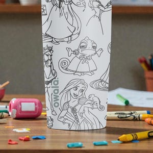 May include: White tumbler featuring line art of cartoon characters. The tumbler has a variety of characters, including a princess, a horse, and a chameleon. The brand name "owala" is printed on the side. The tumbler is surrounded by crayons and other art supplies.