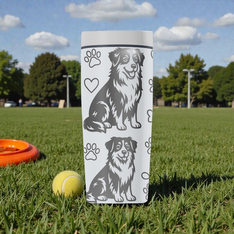 Puede incluir: Un vaso blanco con una ilustraci&oacute;n gris de un perro pastor australiano, huellas de patas y corazones. El vaso cil&iacute;ndrico est&aacute; sobre hierba. Una pelota de tenis y un frisbee est&aacute;n en primer plano. El vaso mide unos 20 cm de alto.