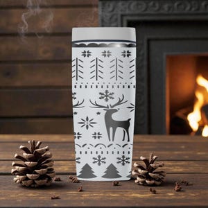 May include: A white travel mug with a black and gray winter pattern featuring trees, snowflakes, and a reindeer. Steam rises from the top of the mug. The mug is on a wooden surface with pine cones.