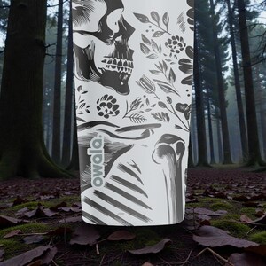 May include: A white Owala water bottle featuring a black skull and floral design. The bottle has a black and white skeletal illustration and the Owala logo. The background is a forest scene with tall trees and fallen leaves.